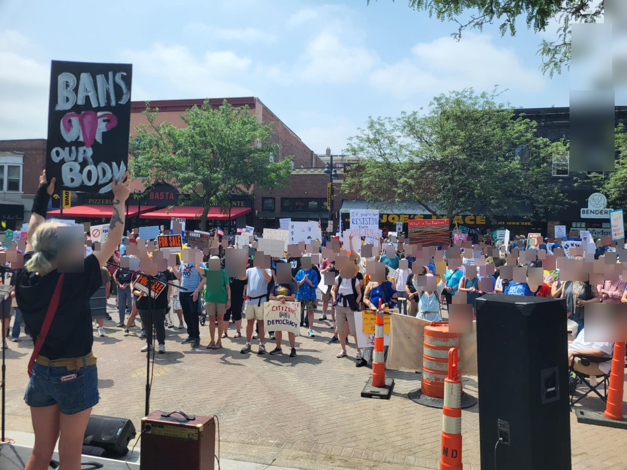 A speaker addresses the crowd at the No Kings Day rally in Iowa City, holding a Bans Off Our Body sign as hundreds of attendees look on
