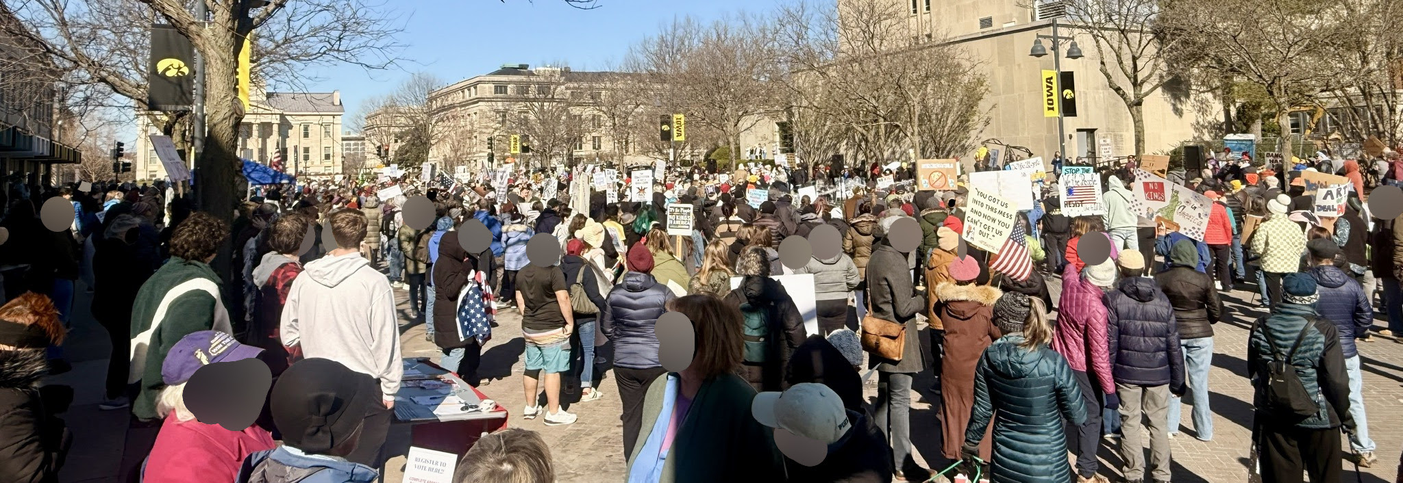 A wide shot of a large crowd of protestors holding signs and listening to a speaker, photographed from behind. Visible faces have been blurred for privacy.