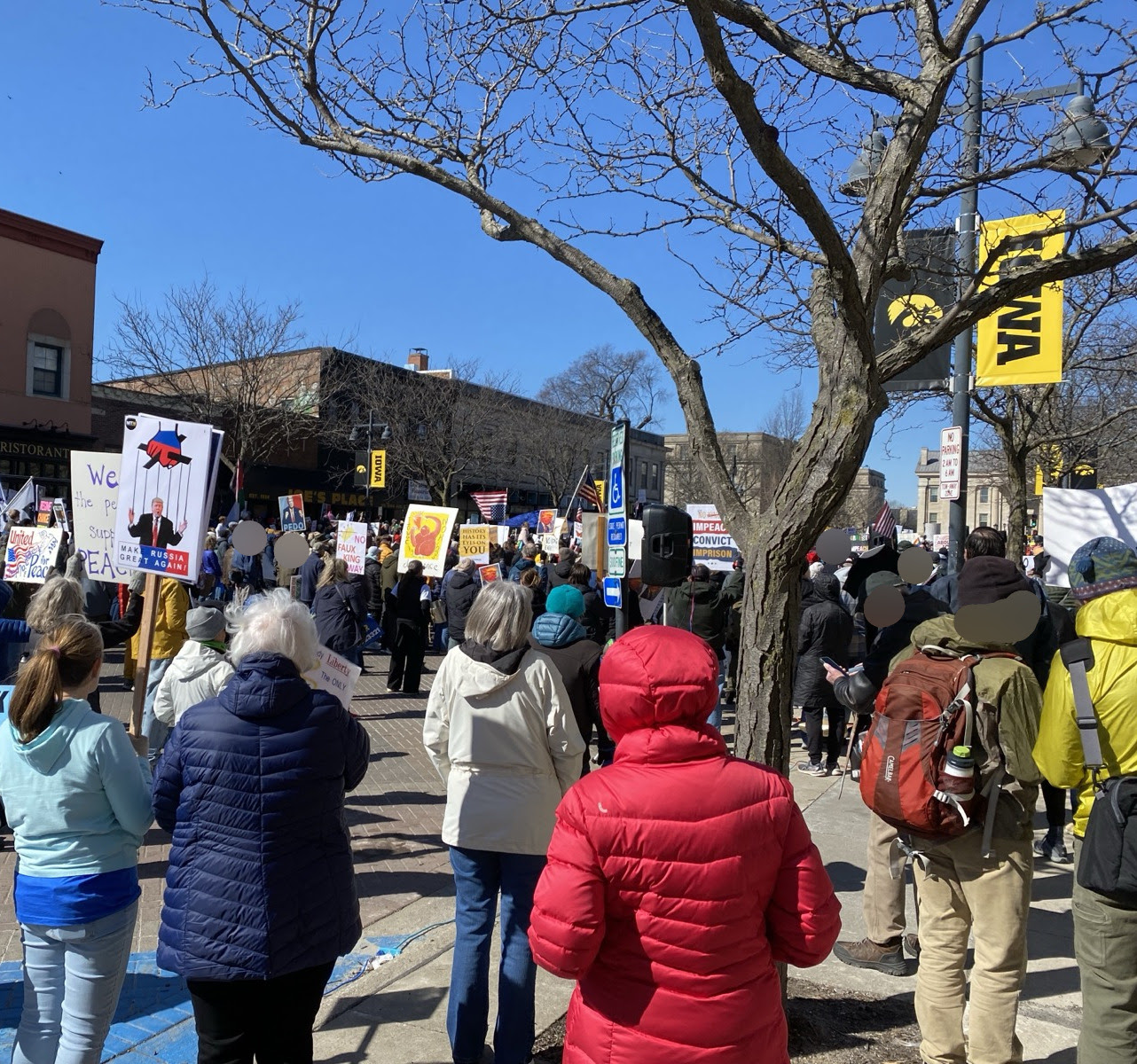 A large crowd of protesters, many holding handmade signs, photographed from behind. Visible faces have been blurred for privacy.
